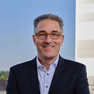 Ein Porträtfoto von Dr. Thomas Obersojer in einem runden Ausschnitt. Er lächelt und trägt eine Brille mit hellem Rahmen, ein dunkles Sakko und ein offenes weißes Hemd. Der Hintergrund ist unscharf und zeigt einen hellblauen Himmel und einen Teil eines weißen Gebäudes.